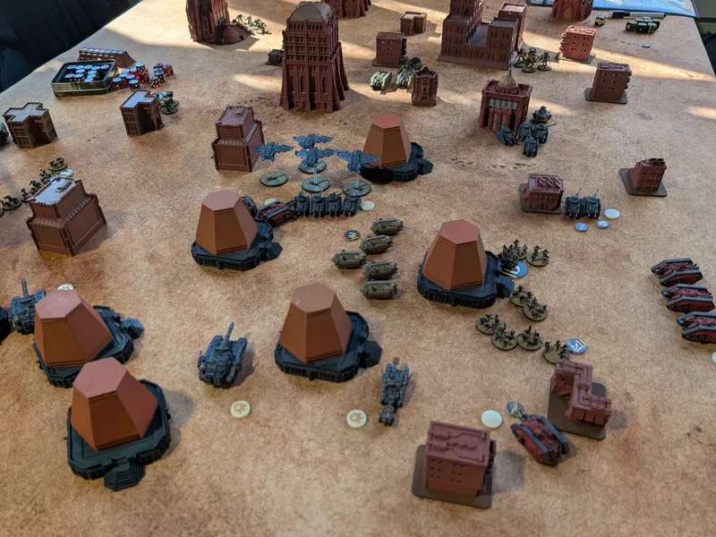 Legions Imperialis tournament table under sunlight: red-primed buildings and terracotta bunkers with grey and red tank squadrons and a flight of aircraft between them.