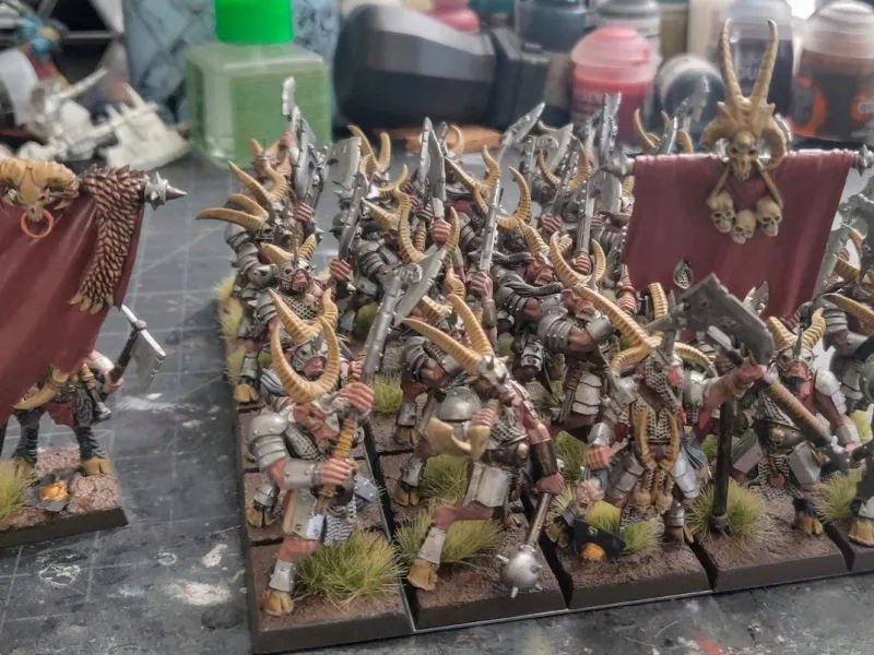 A regiment of horned Chaos warriors with red banners, led by a standard bearer, on a paint-spattered workbench.