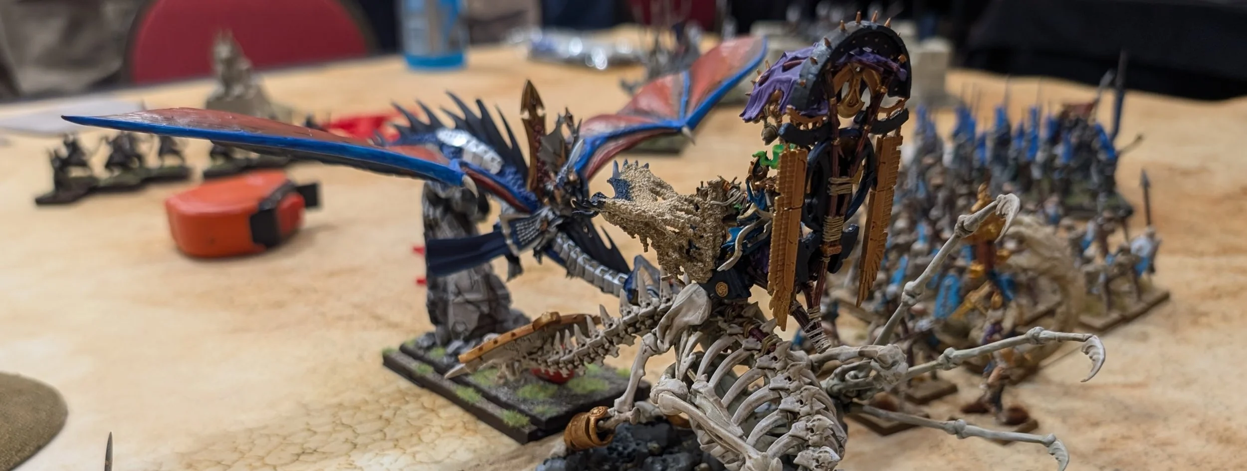Close-up of a tournament table: a skeletal bone dragon with a howdah in the foreground, a blue-winged dragon behind it, and ranked blue infantry off to the right.