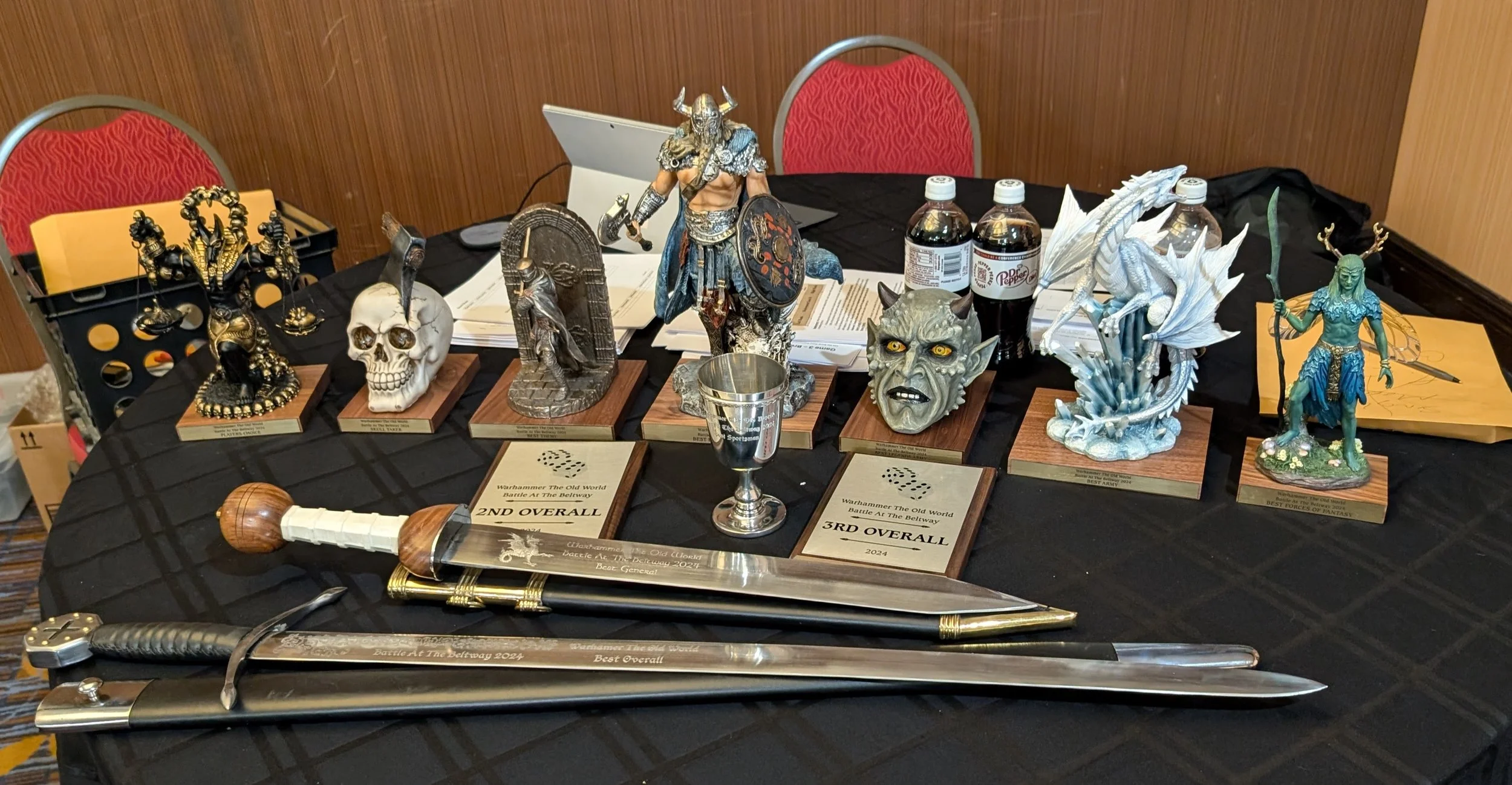 A table of tournament prizes: two engraved swords, a pewter goblet, and a row of resin statue trophies on wooden plaques.