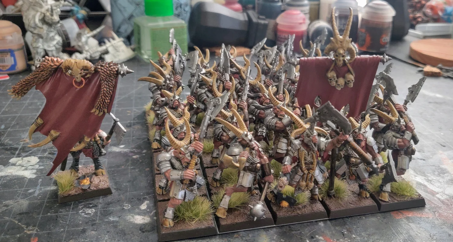 A regiment of horned Chaos warriors with red banners, led by a standard bearer, on a paint-spattered workbench.