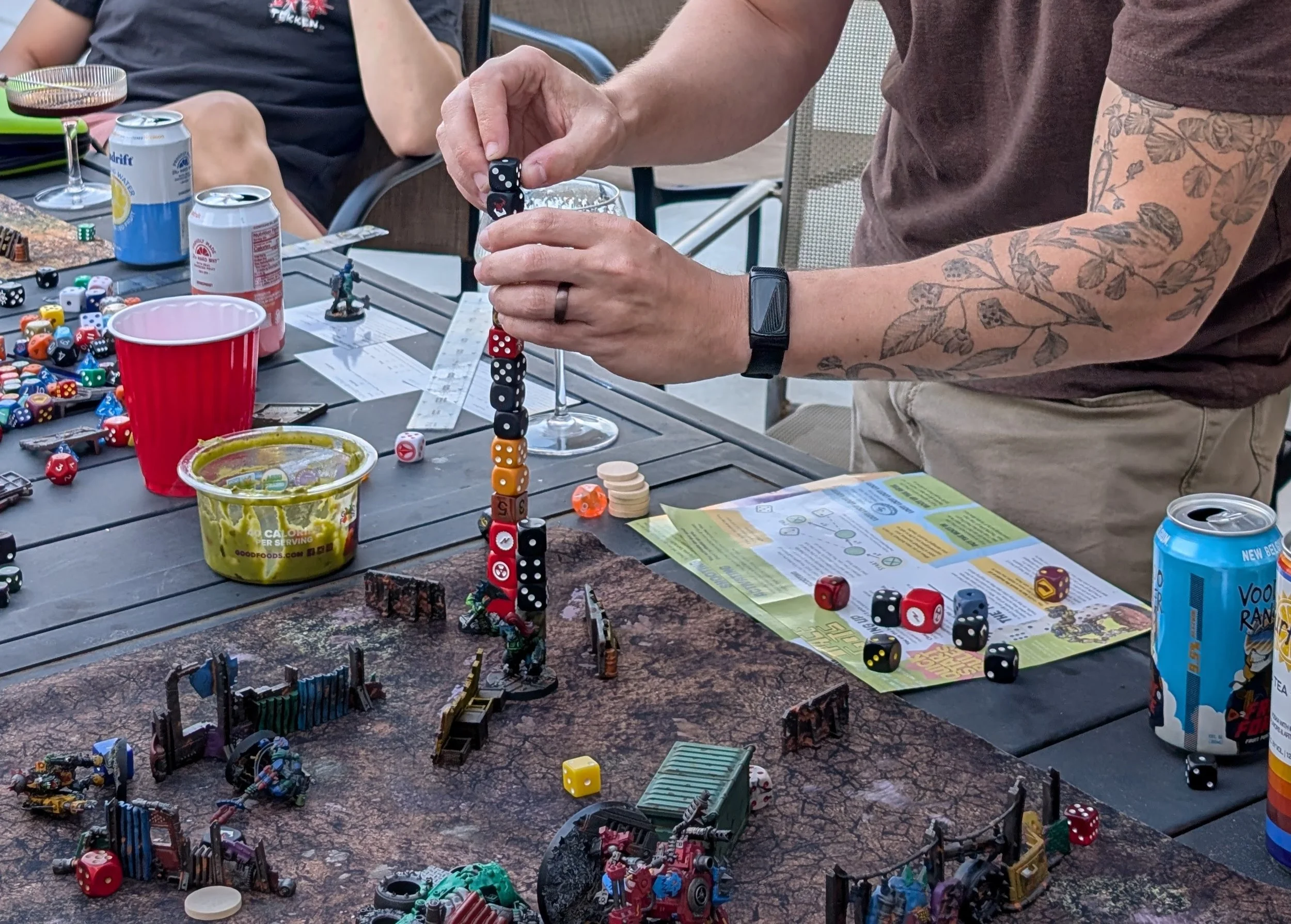 A player carefully stacking a column of dice above an ork model on a patio table crowded with drinks, guacamole, rules sheets, and scatter terrain.