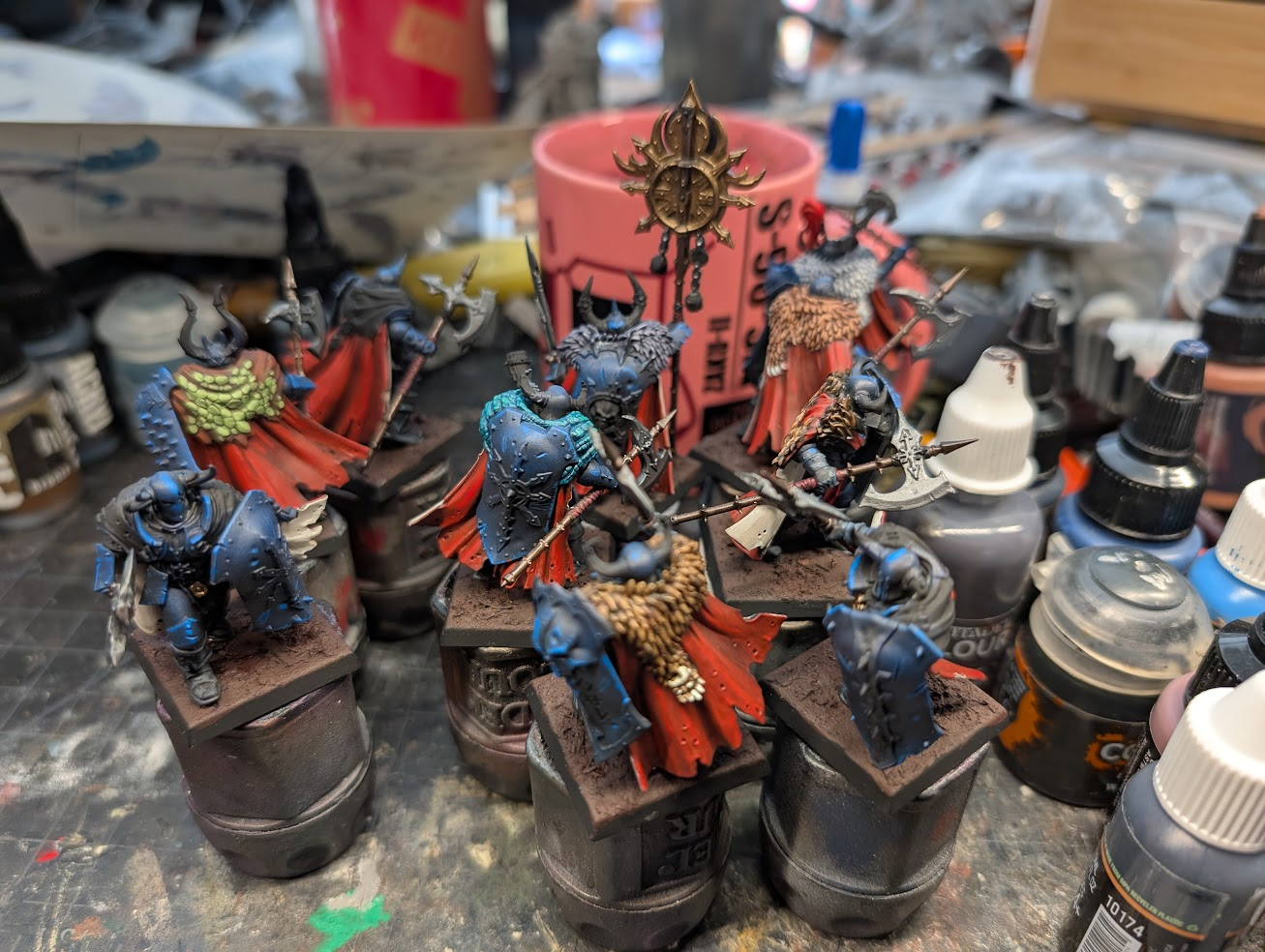 Six part-painted Warriors of Chaos on paint-pot handles, blue armour and red cloaks, surrounded by paint bottles on a cluttered desk.