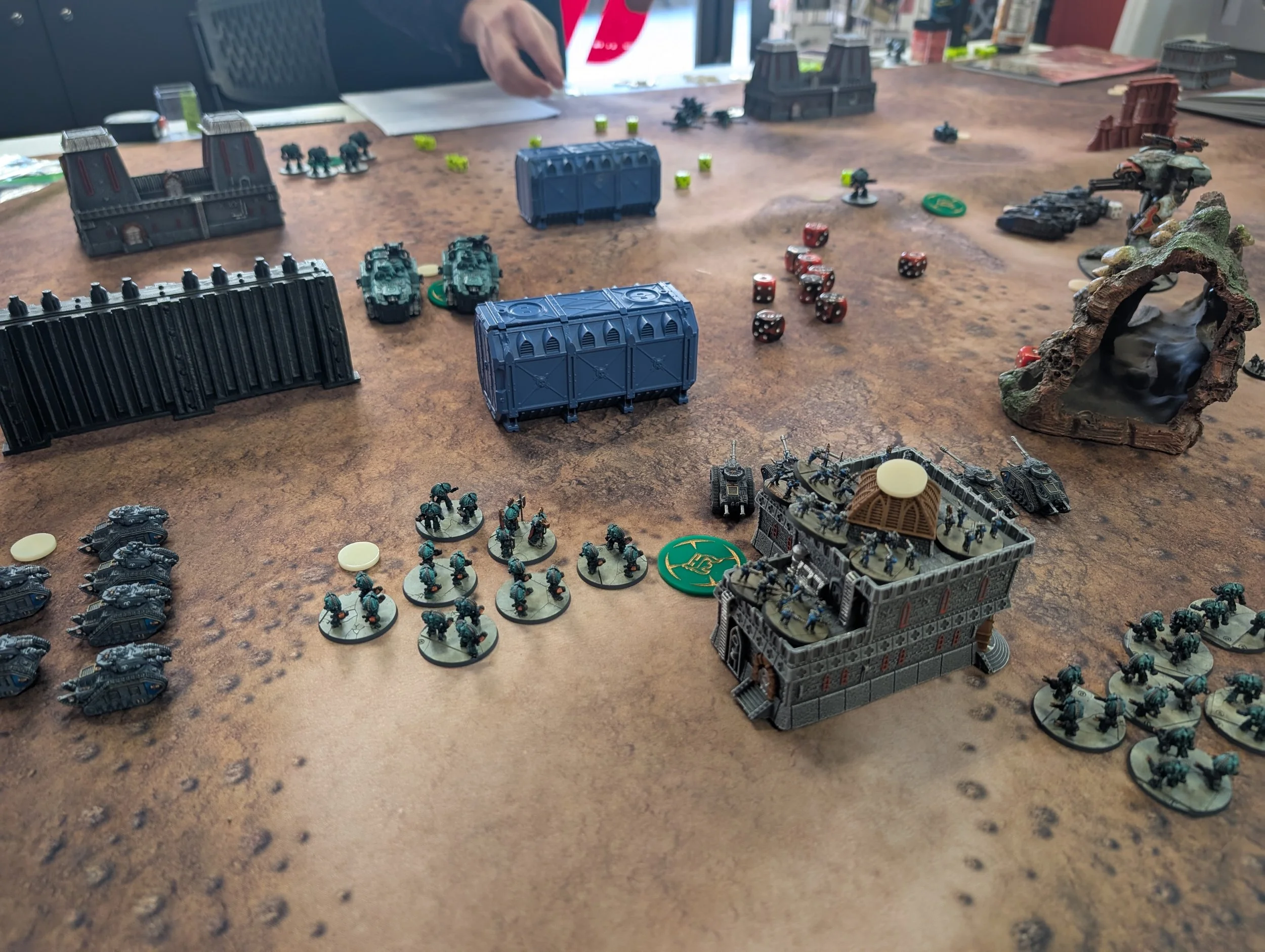 Legions Imperialis table on a rust-coloured mat: green Sons of Horus infantry stands and tanks in the foreground, blue cargo containers and gothic buildings as terrain.