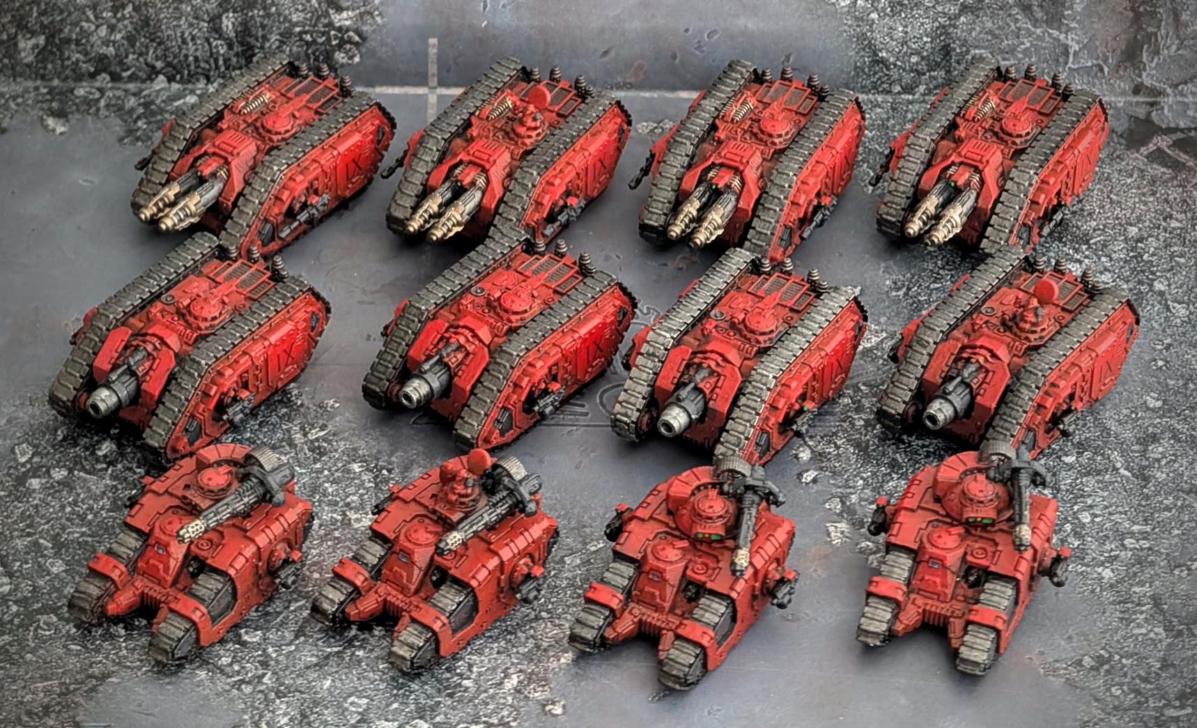 Twelve freshly painted red Blood Angels tanks in three rows on a grey work surface.