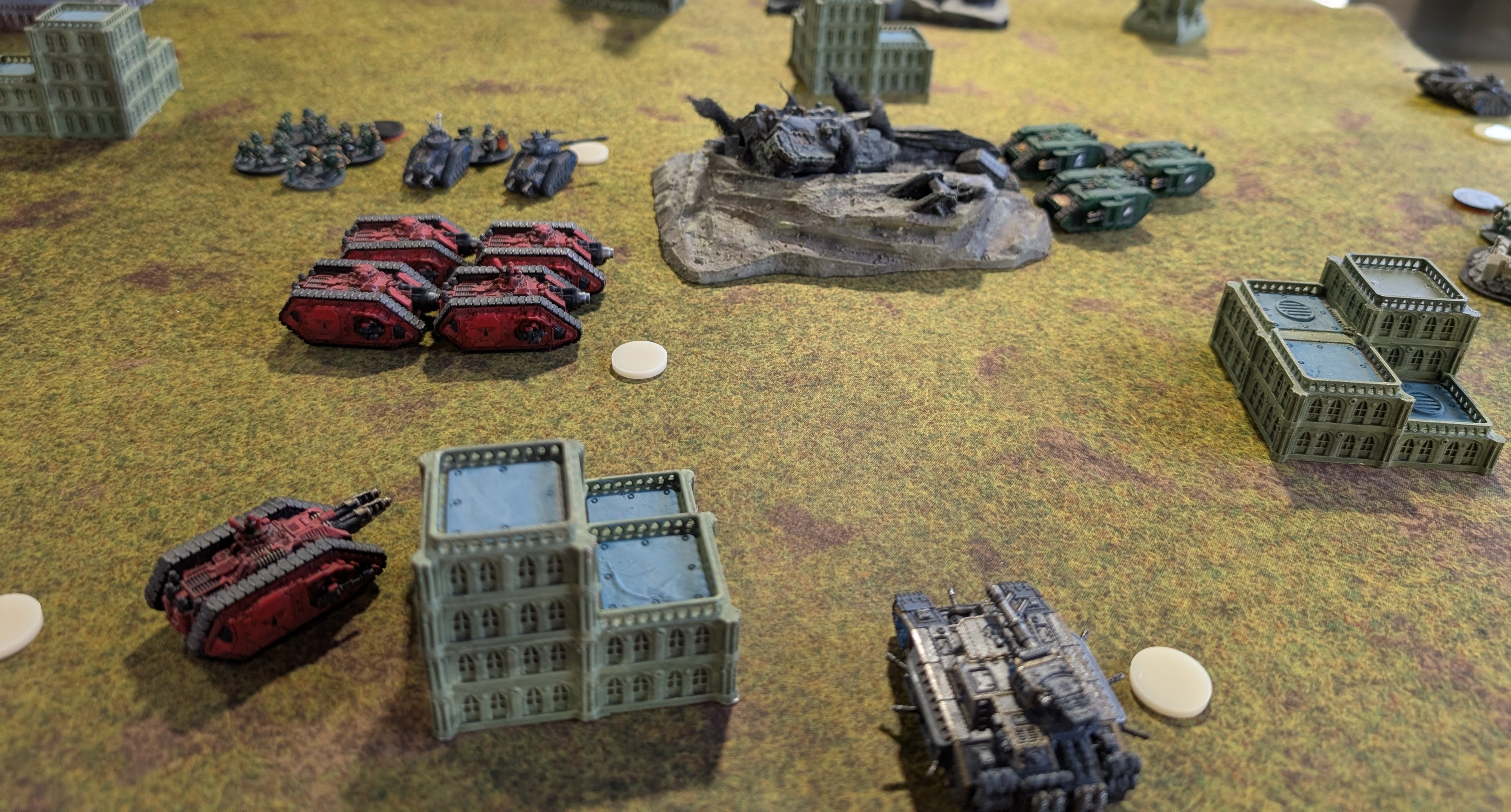Legions Imperialis event table: red Blood Angels tanks facing green and grey vehicles across a yellow-green mat, a wrecked tank on a rocky outcrop between them.