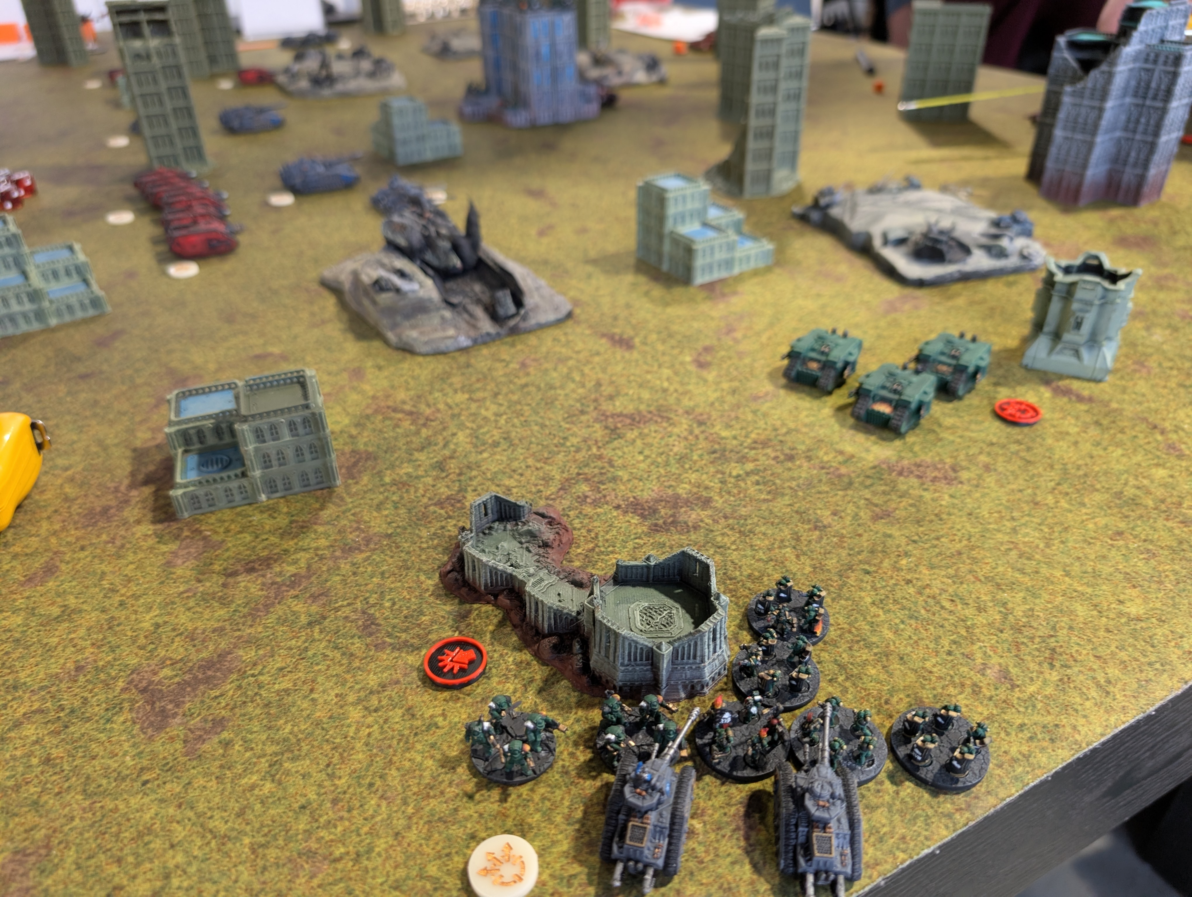 Epic-scale Legions Imperialis game in progress: green infantry stands and grey tanks holding gothic ruins on a mossy battlefield, red tanks advancing at the far side.