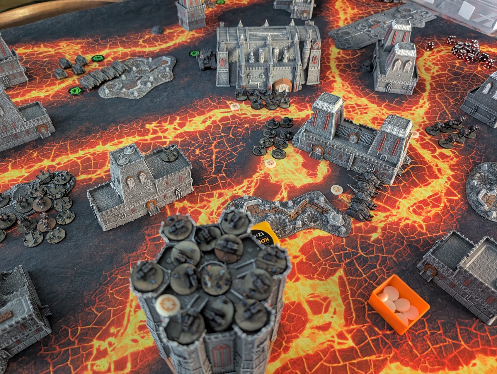 Legions Imperialis game on a lava-cracked mat: grey gothic buildings scattered across the board with epic-scale infantry stands and tanks between them.